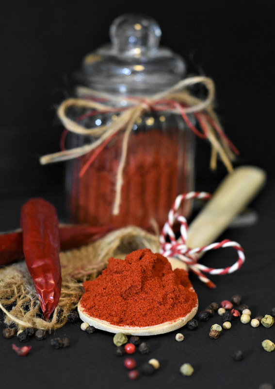 Wooden spoon tied with red and white string and filled with reddish-orange paprika, dried peppercorns, a strip of coarse macrame and two dried red peppers in the foreground, a glass jar filled with paprika and tied with natural and red raffia bow midground on a black background